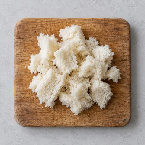 Crustless day-old white bread torn into pieces on a wooden cutting board.