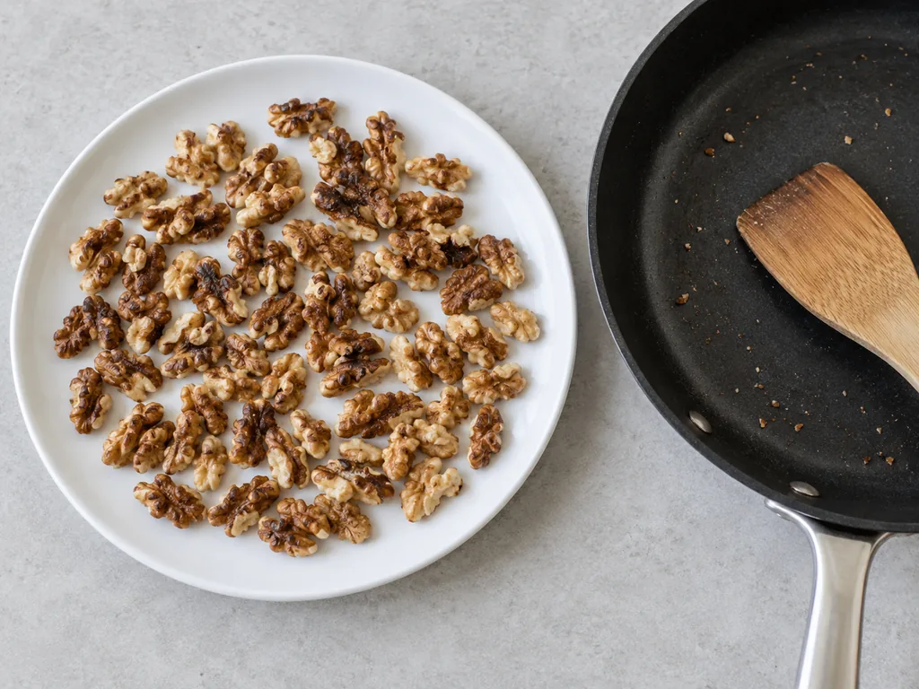 Toasted walnut pieces cool in a single layer on a white plate beside the empty skillet.