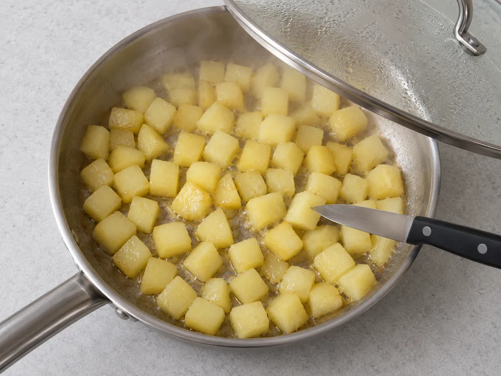 Pale potato cubes steam beneath a lifted skillet lid while a knife tests one cube.