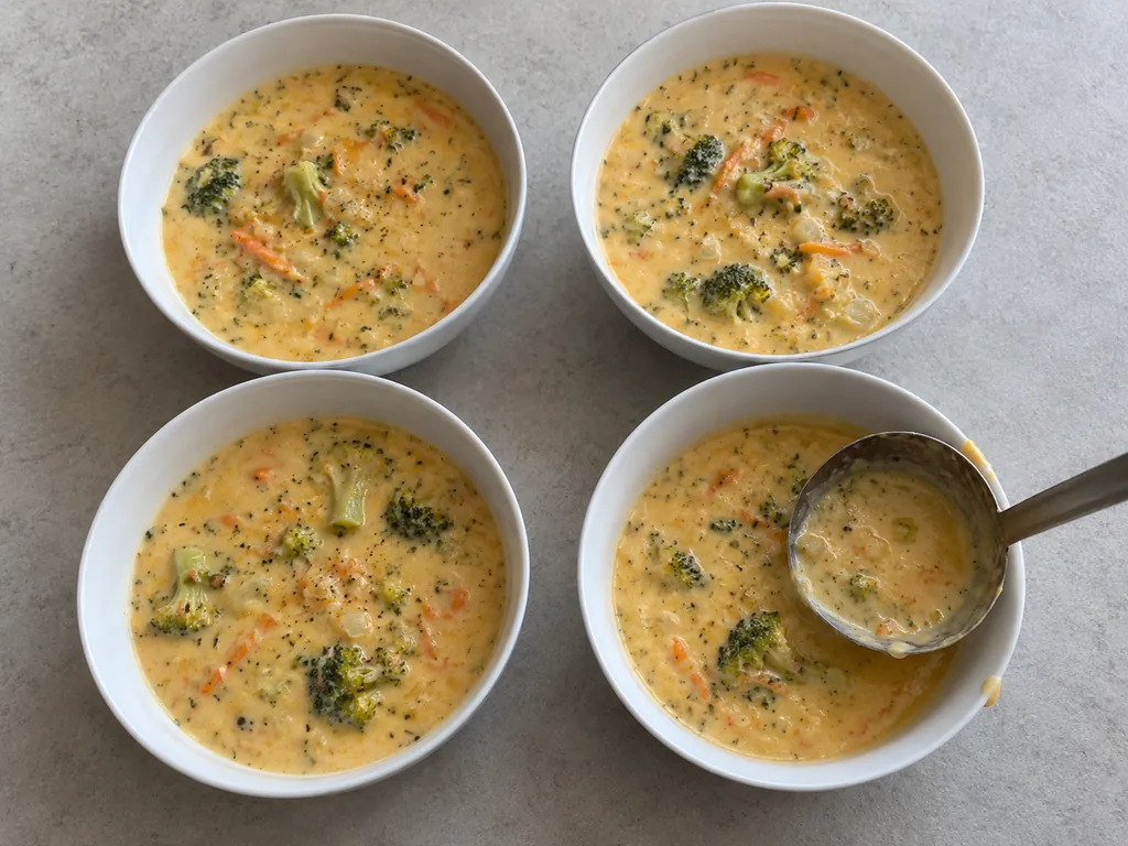 Four white bowls hold creamy broccoli cheddar soup finished with scattered black pepper.