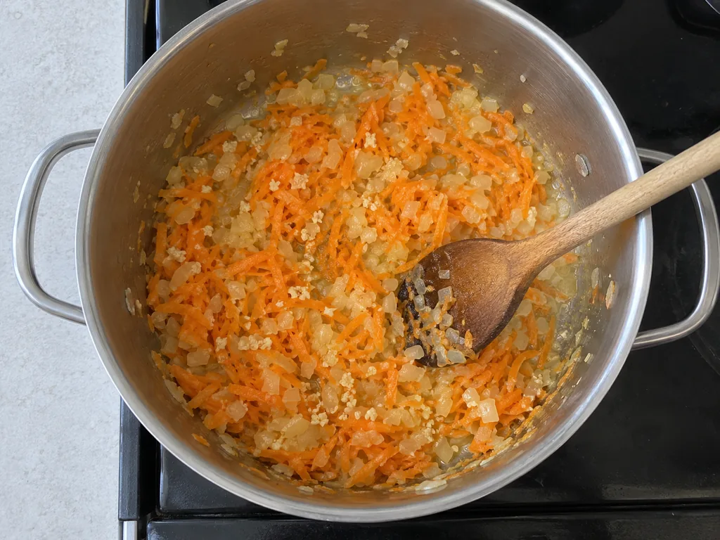 Minced garlic flecks mingle with softened onion and carrot beside a wooden spoon.