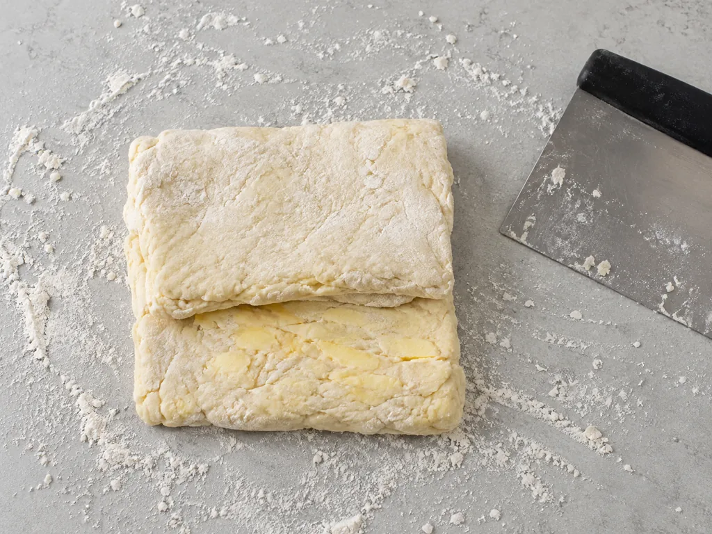 Biscuit dough folds over itself on a floured counter beside a bench scraper.