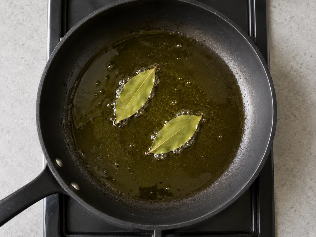 Two bay leaves sizzle in shimmering olive oil inside a black skillet.