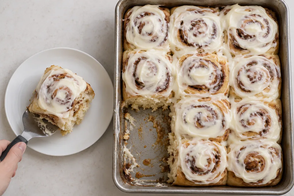 A spatula transfers one frosted cinnamon roll to a plate, revealing tender crumb beside the remaining pan.