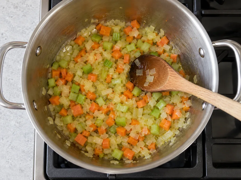 Translucent onion, diced carrot, and celery glisten in olive oil around a wooden spoon.