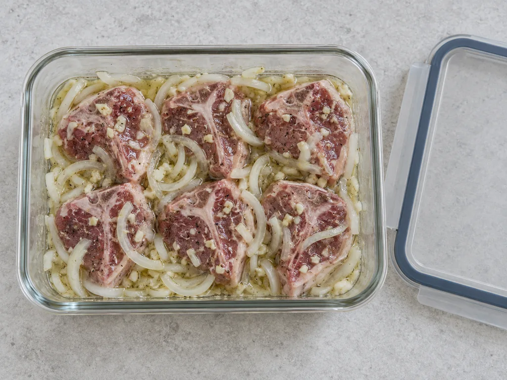 Lamb loin chops are coated with onion marinade inside a glass container beside its lid.