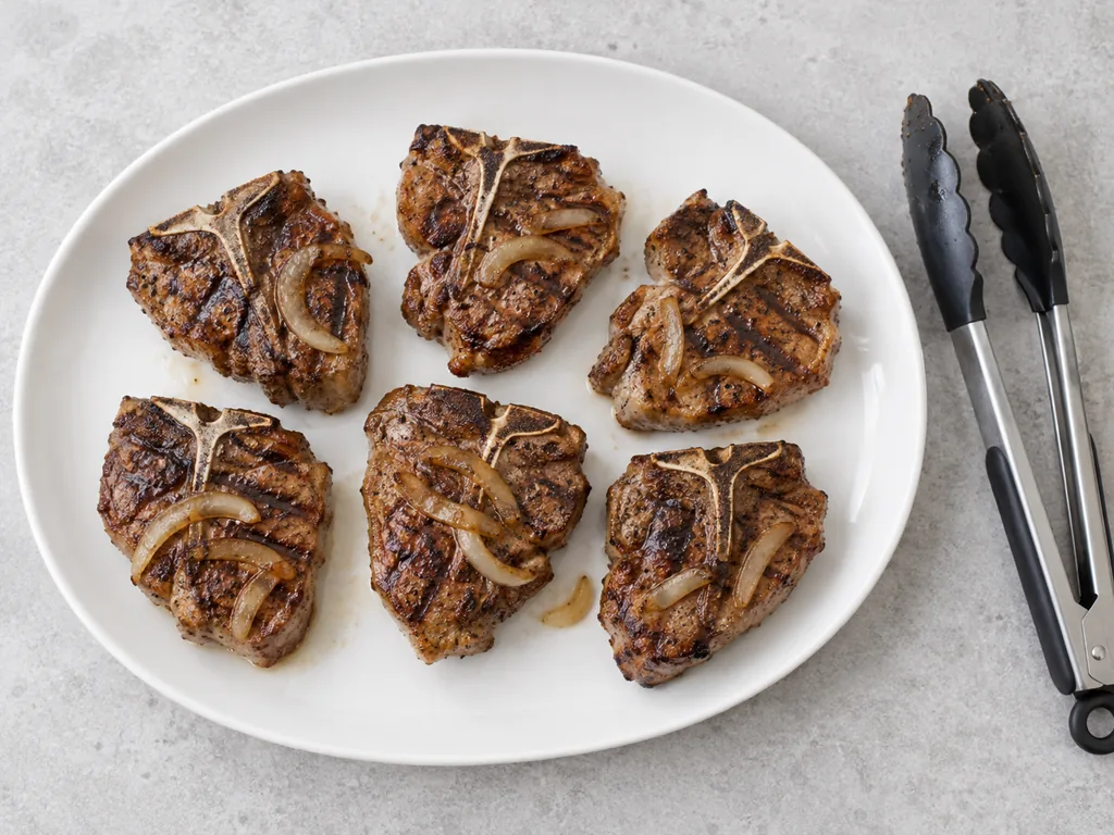 Six grilled lamb loin chops rest on a clean white platter beside tongs.