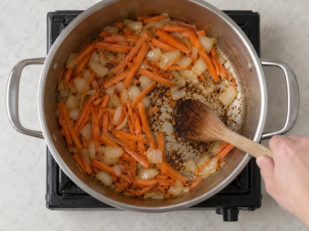 A wooden spoon scrapes browned fond beneath softening onion pieces and thin carrot strips in the pot.